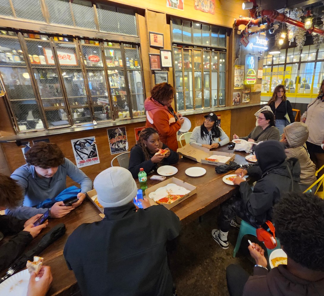 a group of students sitting at a table eating pizza.